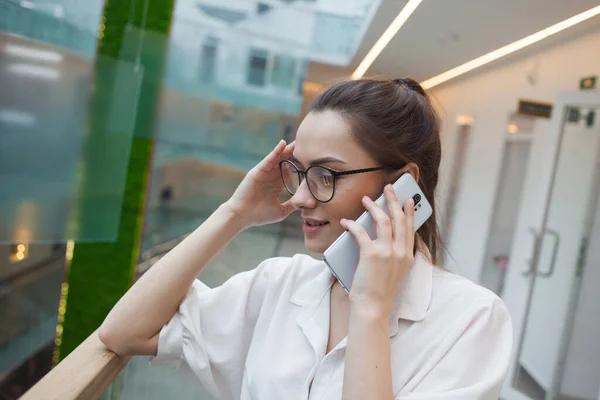 Conversación telefónica, una joven mujer emocional con una camisa blanca y gafas hablando en un teléfono móvil — Foto de Stock