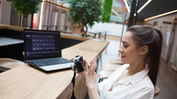 young attractive girl is sitting in front of a laptop and holding a camera in her hands. Photographer and Retoucher