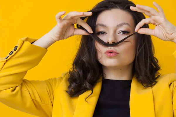 Stilvolle junge Frau in gelber Jacke vor gelbem Hintergrund, die sich einen Schnurrbart aus den Haaren zieht. — Stockfoto