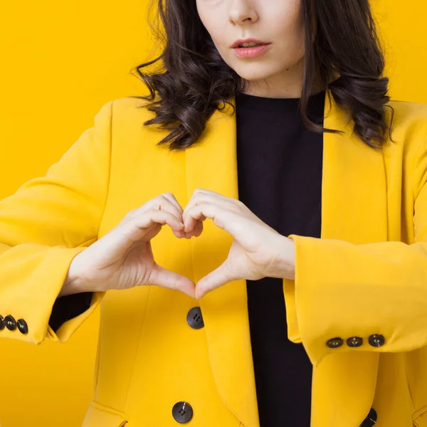 Eine junge brünette Frau in gelbem Sakko zeigt eine Geste des Herzens. Ein Zeichen der Liebe und Unterstützung. — Stockfoto