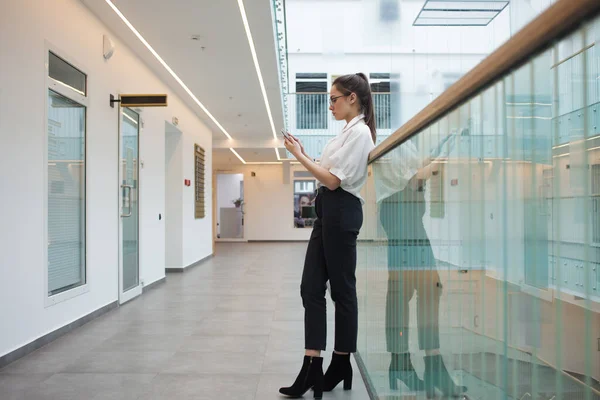 En ung positiv affärskvinna i glasögon och en vit skjorta använder en smartphone för att kommunicera med kollegor, — Stockfoto