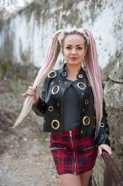 A girl with dreadlocks in a leather jacket and a short skirt stands against the background of an old stone wall — Stock Photo, Image