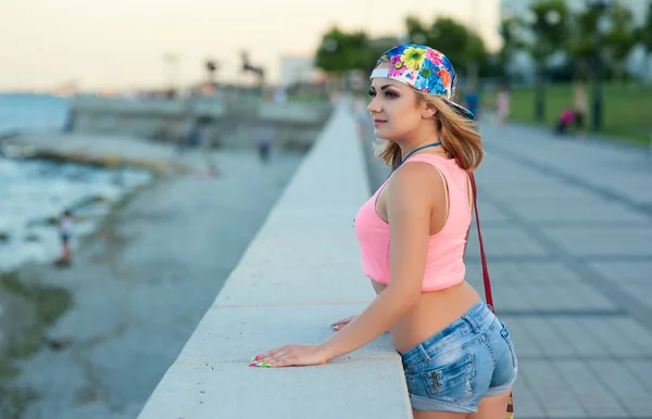Sexy jovem loira menina no denim shorts stands no cidade beira-mar e olha para o mar — Fotografia de Stock