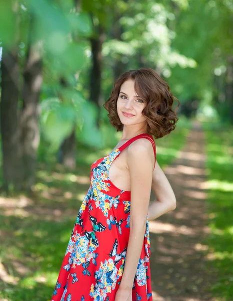 Jovem morena em vestido vermelho posando no beco no parque de verão contra árvores — Fotografia de Stock