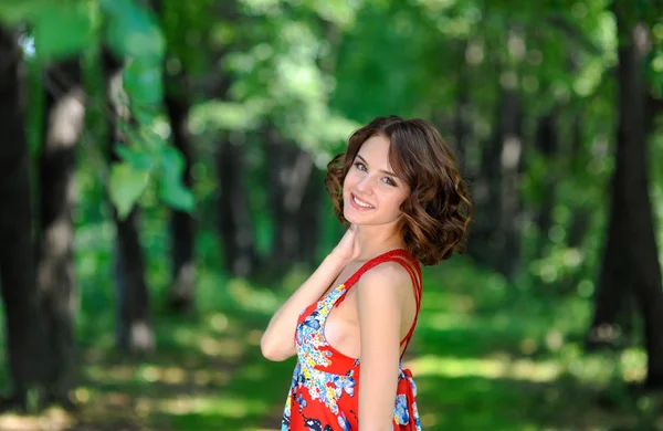 Jeune fille brune en robe rouge posant sur l'allée dans le parc d'été contre les arbres — Photo