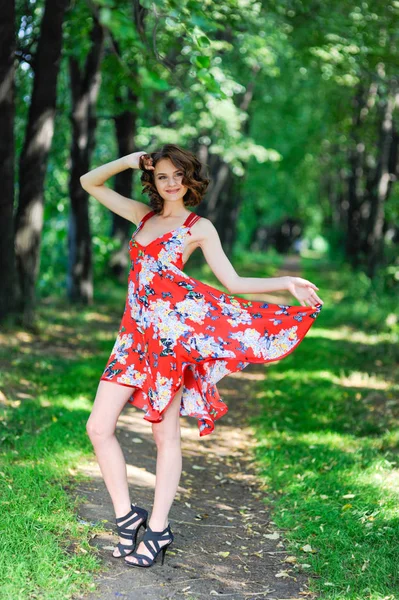 Une jeune fille brune en robe rouge danse sur une avenue dans un parc d'été sur fond d'arbres — Photo