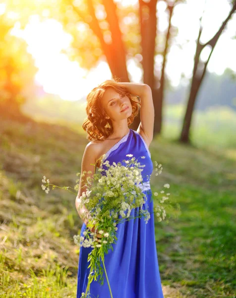 Молодая брюнетка в синем платье, позирующая с букетом диких цветов в парке в лучах яркого солнца — стоковое фото