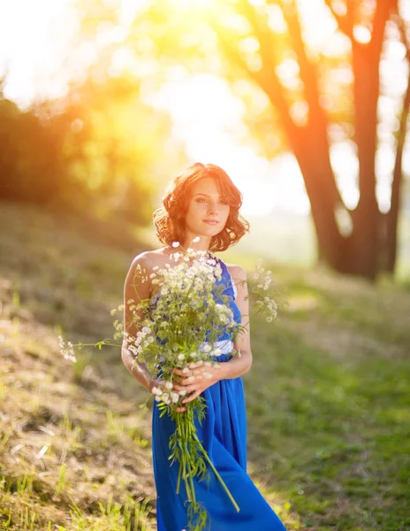 Una giovane ragazza bruna in abito blu in posa con un mazzo di fiori selvatici in un parco ai raggi di un sole luminoso — Foto Stock