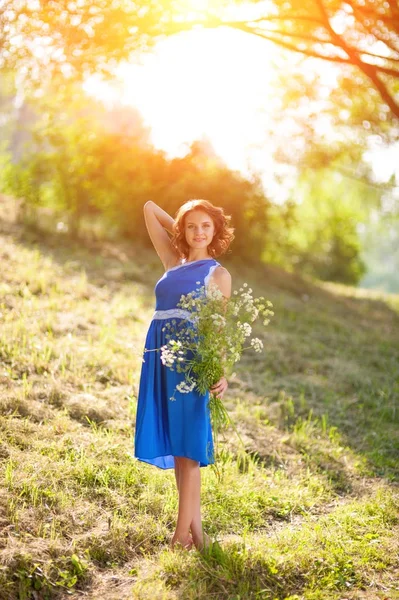 Ein junges brünettes Mädchen in einem blauen Kleid posiert mit einem Strauß wilder Blumen in einem Park in den Strahlen einer hellen Sonne — Stockfoto