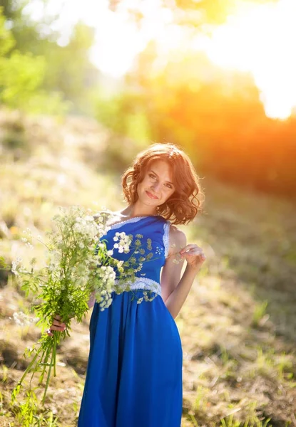 Egy fiatal barna lány egy kék ruhás pózol egy-egy csokor a vadvirágok, egy parkban a fényes nap sugarai — Stock Fotó