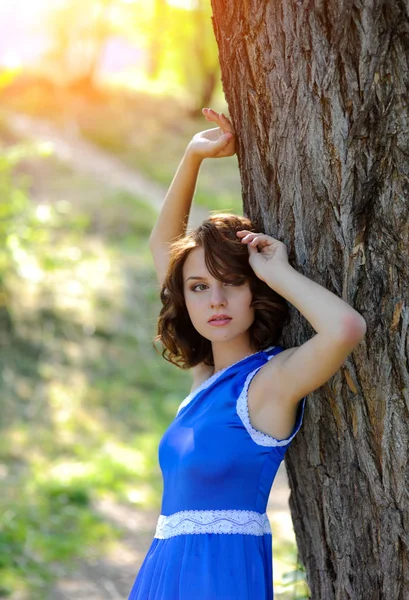 Mladá bruneta dívka v modrých šatech představuje v blízkosti velký strom v letním parku v paprscích slunce — Stock fotografie