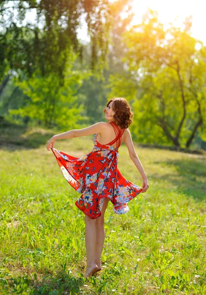 Junges brünettes Mädchen in rotem Kleid posiert auf Gras im Sommerpark in der Sonne — Stockfoto