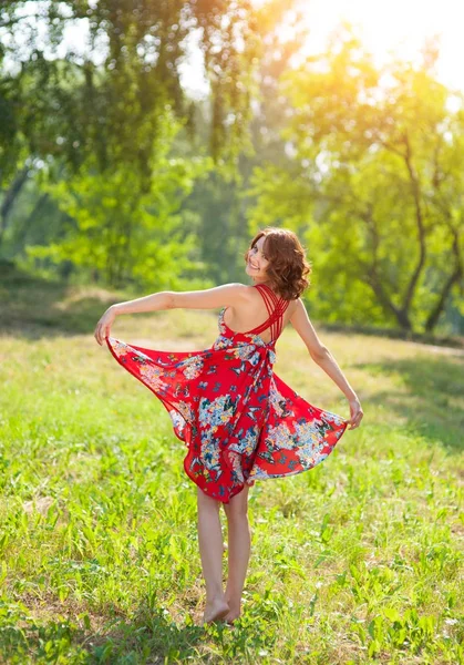 Junges brünettes Mädchen in rotem Kleid posiert auf Gras im Sommerpark in der Sonne — Stockfoto