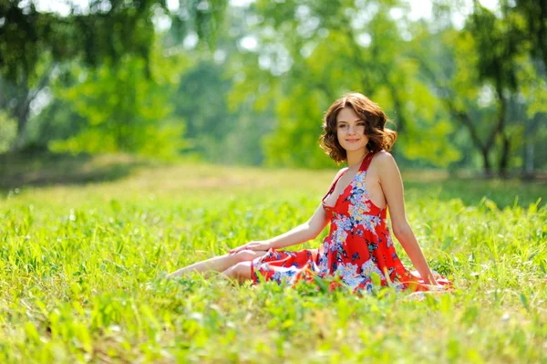 Jeune fille brune en robe rouge posant sur l'herbe dans le parc d'été au soleil — Photo
