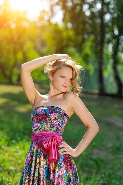 Молода блондинка в яскравій барвистій сукні позує в літньому парку в променях яскравого сонця — стокове фото