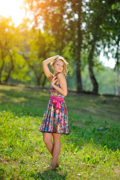 Menina loira jovem em vestido colorido brilhante posando em um parque de verão nos raios de um sol brilhante — Fotografia de Stock