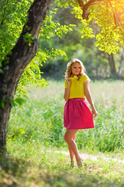 Menina loira nova em uma blusa amarela com uma saia rosa brilhante posando em um parque de verão nos raios de um sol brilhante — Fotografia de Stock