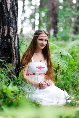 Güzel, genç bir Slav kız uzun saç ve Slav etnik kıyafetleri ile bir yaz ormanda oturur ve bir meyve onun elinde tutan
