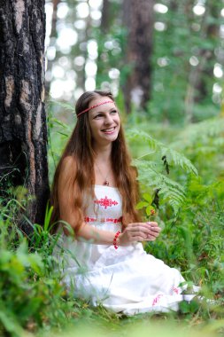 Güzel, genç bir Slav kız uzun saç ve Slav etnik kıyafetleri ile bir yaz ormanda oturur ve bir meyve onun elinde tutan