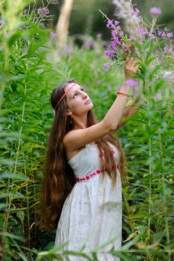 Uzun saçlı ve Slav etnik elbise genç güzel bir Slav kız yaz ormanda uzun boylu çalılar arasında duruyor ve onun elinde bir çiçek tutan