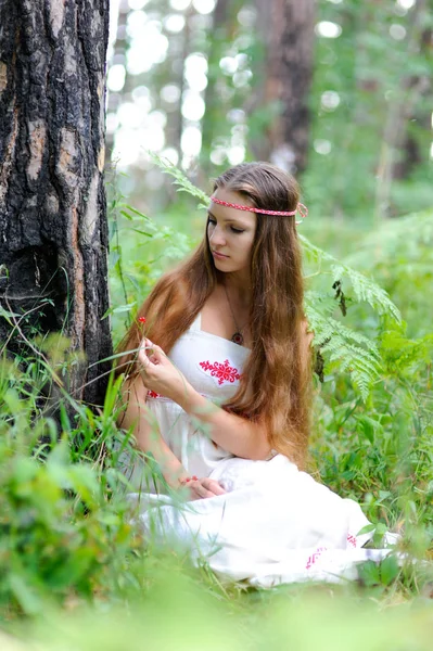 Uma menina eslava bonita nova com cabelo longo e traje étnico eslavo senta-se em uma floresta de verão e segura uma baga em suas mãos — Fotografia de Stock