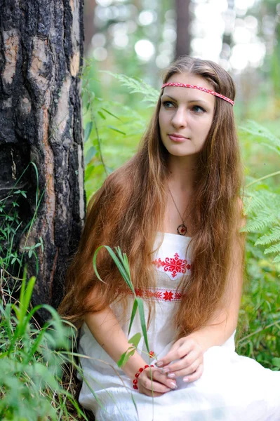 Молодая красивая славянская девушка с длинными волосами и славянской этнической одеждой сидит в летнем лесу и держит в руках ягоду — стоковое фото