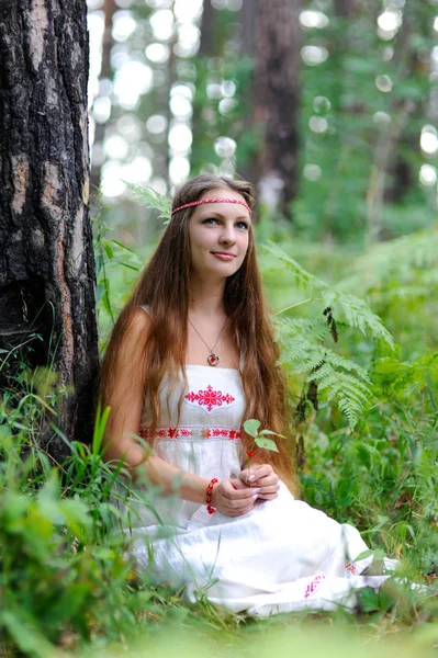 A young beautiful Slavic girl with long hair and Slavic ethnic attire sits in a summer forest and holds a berry in her hands — Stock Photo, Image