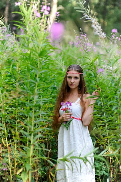 Une jeune belle fille slave aux cheveux longs et à la robe ethnique slave se tient parmi les hauts sous-bois de la forêt d'été et tient une fleur dans ses mains — Photo