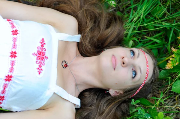 Menina eslava bonita nova com cabelos longos e trajes étnicos eslavos encontra-se na grama em uma floresta de verão — Fotografia de Stock