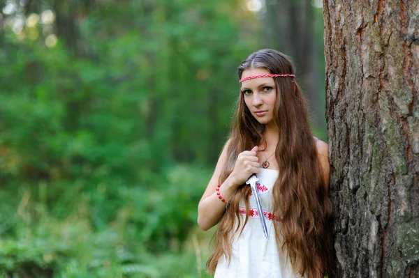 Uma menina eslava bonita nova com cabelo longo e vestido étnico eslavo está em uma floresta de verão com uma adaga ritual em suas mãos — Fotografia de Stock