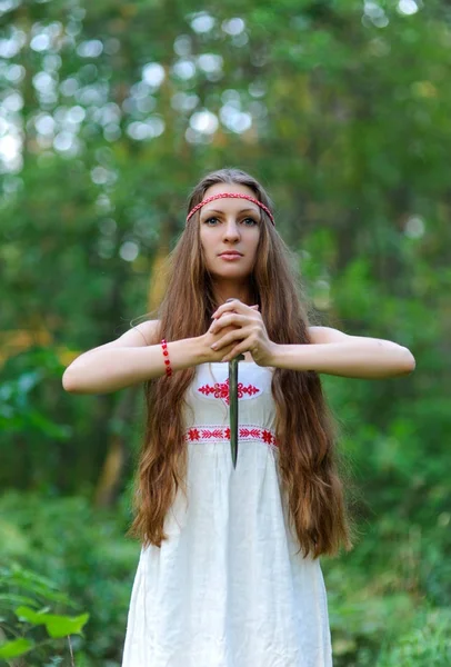 Krásná mladá Slovanská dívka s dlouhými vlasy a slovanskou etnickou šaty stojí v lese letní s rituální dýku v ruce — Stock fotografie