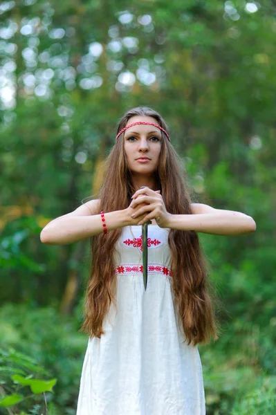 Uma menina eslava bonita nova com cabelo longo e vestido étnico eslavo está em uma floresta de verão com uma adaga ritual em suas mãos — Fotografia de Stock