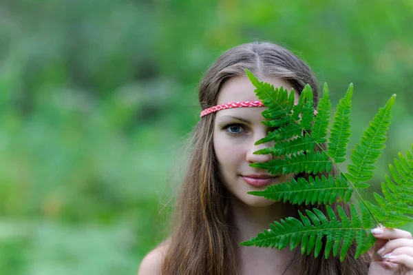 A young beautiful Slavic girl with long hair and Slavic ethnic dress covered her face with a fern leaf — Stock Photo, Image