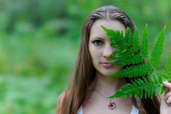 Une jeune belle fille slave aux cheveux longs et à la robe ethnique slave a recouvert son visage d'une feuille de fougère — Photo