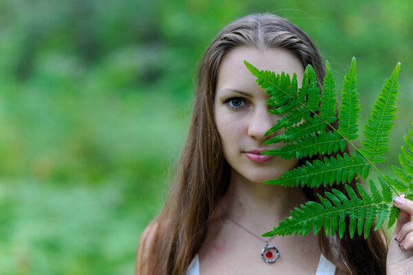 A young beautiful Slavic girl with long hair and Slavic ethnic dress covered her face with a fern leaf