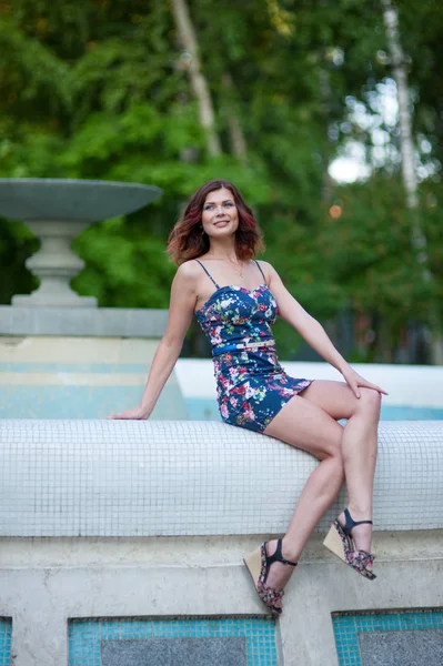 Kısa bir elbise içinde dalgalı saçlı güzel genç esmer kadın parkta bir çeşme kenarında oturur