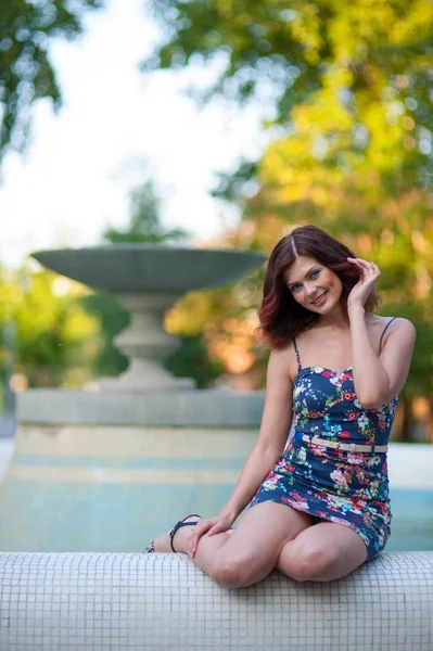 Kısa bir elbise içinde dalgalı saçlı güzel genç esmer kadın parkta bir çeşme kenarında oturur
