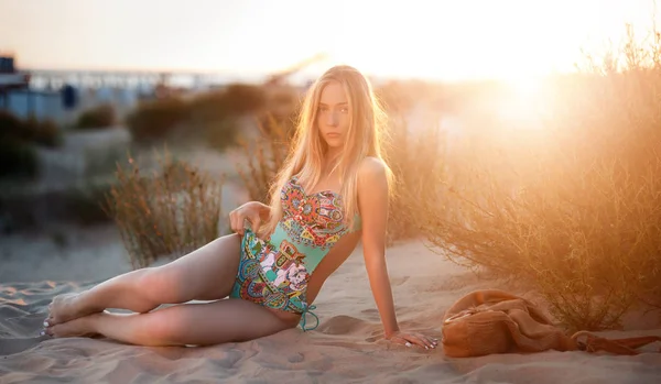 Sexy blonde in a bathing suit lying on the sand at sunset in the rays of a bright sun — Stock Photo, Image