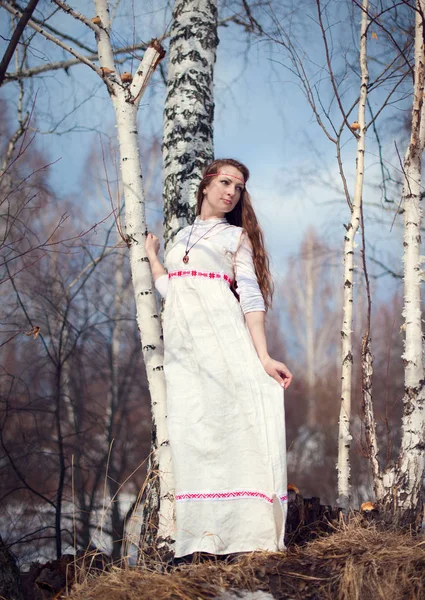 Young beautiful Slavic girl with long hair and Slavic ethnic attire posing in spring forest near birch — Stock Photo, Image