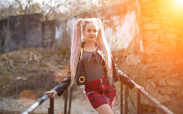 Uma menina com dreadlocks em uma jaqueta de couro e uma saia curta fica contra o fundo de uma parede de pedra velha nos raios de um sol brilhante — Fotografia de Stock