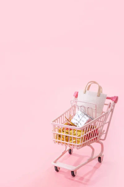 Konsep tahunan musim belanja - trem keranjang mini berwarna merah muda yang penuh dengan kantong kertas yang terisolasi dengan latar belakang berwarna merah muda pucat, ruang fotokopi kosong, tutup — Stok Foto
