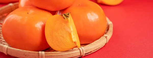 Frische schöne in Scheiben geschnittene süße Persimmon Kaki isoliert auf rotem Tischhintergrund und Bambussieb, chinesisches Mondneujahr Designkonzept, Nahaufnahme. — Stockfoto