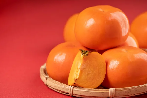 Fresh beautiful sliced sweet persimmon kaki isolated on red table background and bamboo sieve, Chinese lunar new year design concept, close up. — Stock Photo, Image