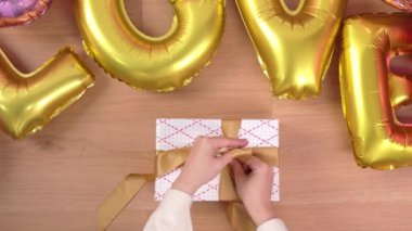Beyaz süveter giyen genç bir kadın hediye paketini sarıp altın balonla tatil hazırlamak için paketliyor. Yay düğümü, yaşam tarzı ve üst görünüm bağlamak için ip ve ambalaj kullanıyor..