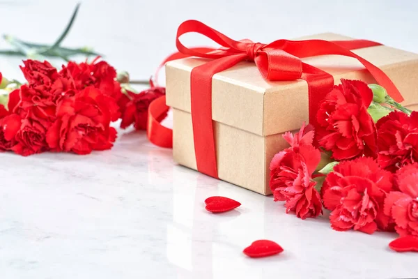 Концепция дизайна ко Дню матери - красивые цветущие красные гвоздики, подарочная коробка с лентой на фоне белого мраморного стола, крупный план, пространство для копирования . — стоковое фото
