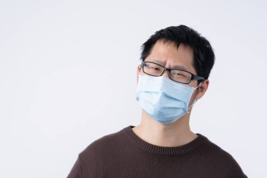 Genç Asyalı erkek portresi, hasta hissediyor, enfeksiyonu önlemek için cerrahi mavi bir maske takıyor, hava kirliliği beyaz arka planda izole edilmiş, kesilmiş..