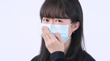 Maskeli bir kadın - genç Asyalı ağzını kapatıyor, beyaz arka planda izole edilmiş tıbbi mavi bir maske takıyor, kendini kötü hissediyor, kapat..