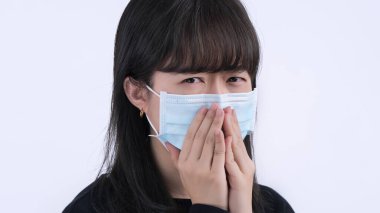 Maskeli bir kadın - genç Asyalı ağzını kapatıyor, beyaz arka planda izole edilmiş tıbbi mavi bir maske takıyor, kendini kötü hissediyor, kapat..