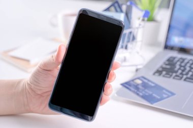 Akıllı cep telefonu taşıyan bir kadın, online ödeme yapma konsepti, beyaz masa arkasında alışveriş yapma konsepti, alışveriş arabası tramvayı, kapat..