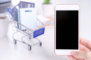 Akıllı cep telefonu taşıyan bir kadın, online ödeme yapma konsepti, beyaz masa arkasında alışveriş yapma konsepti, alışveriş arabası tramvayı, kapat..